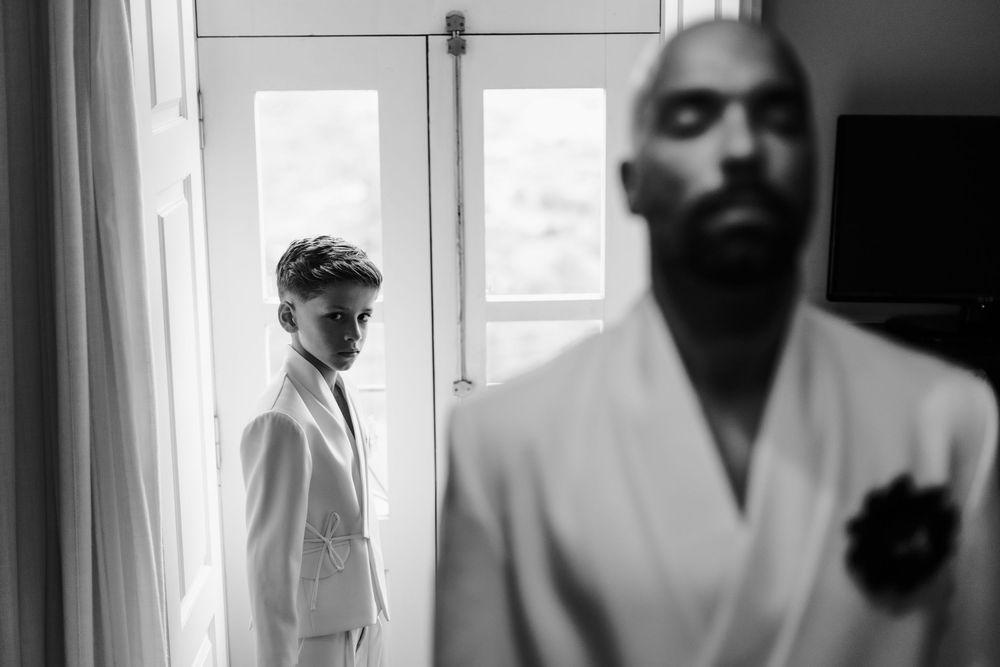 A black and white photo showing two people in white suits standing near a window in an interior setting.