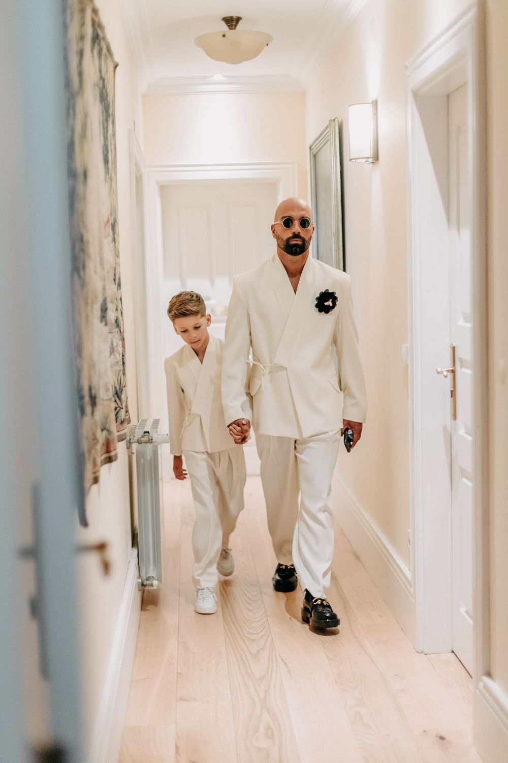 Two people wearing matching all-white outfits walk down a bright hallway with light wood flooring.