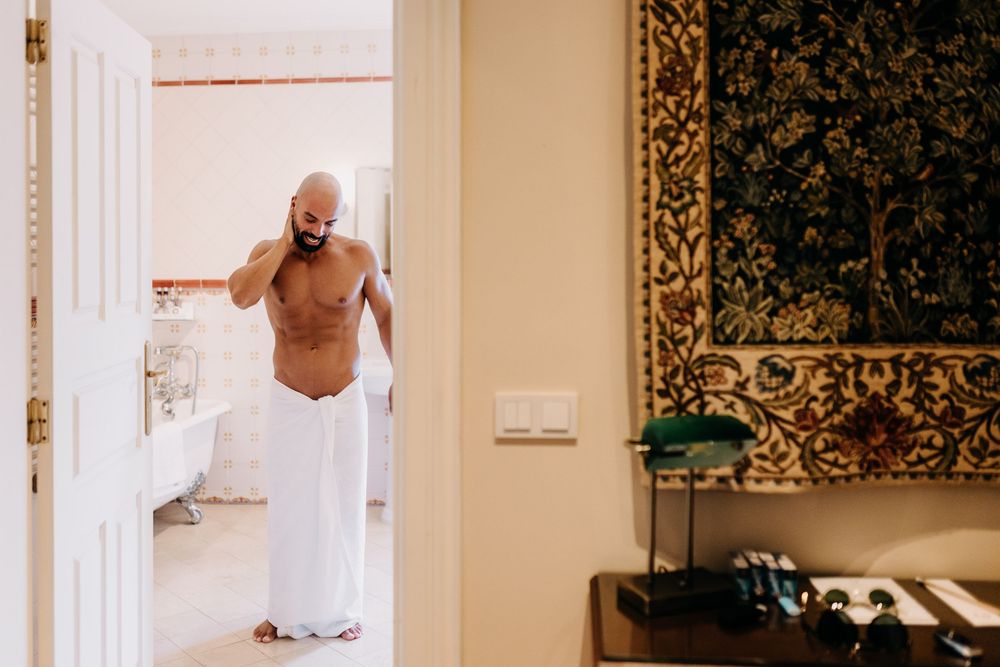 A person wrapped in white fabric peeks through an ornate doorway in an elegant room with a decorative tapestry on the wall.