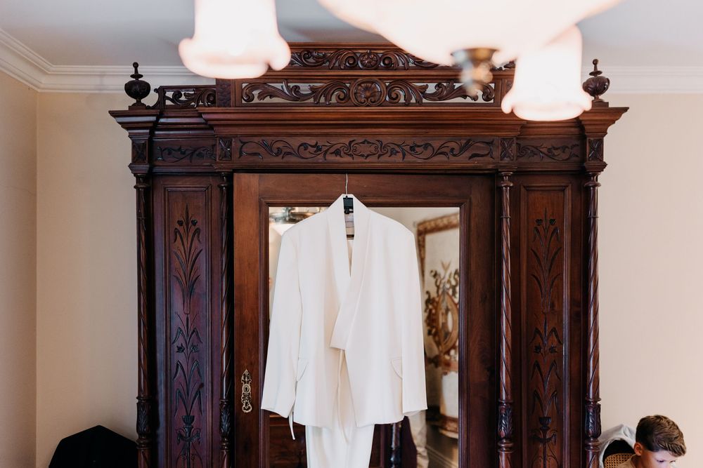 An elegant white wedding suit jacket hangs on an ornate dark wooden vintage armoire with carved details and decorative trim.