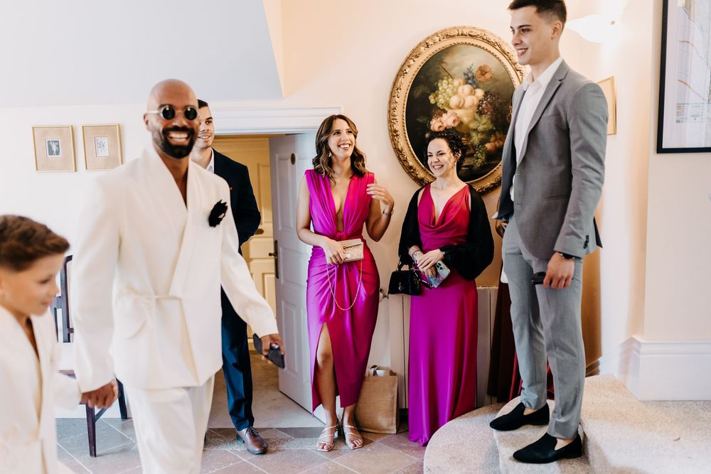 A group in bright formal attire including white and pink dresses with gray suits walk together in an elegant indoor venue.
