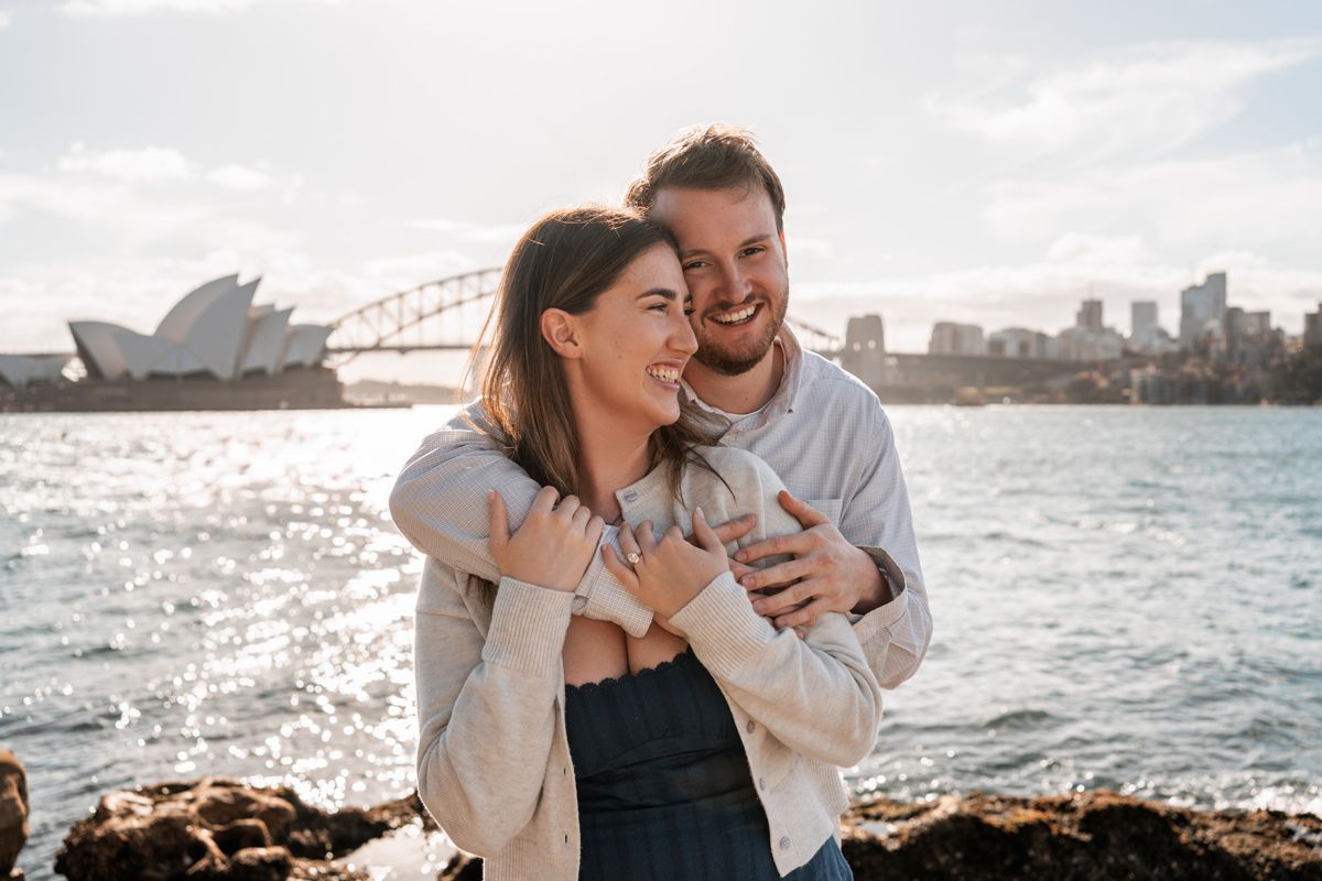 man hugs his girlfriend after a proposal photoshoot in sydney, girl cuddles close to him and both are smiling and are happy