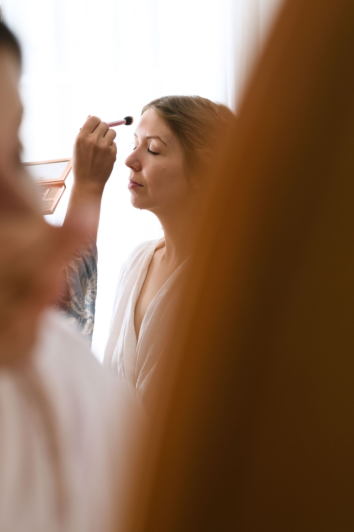 Makeup artist applies eyeshadow on a client wearing a white robe in a soft, naturally lit setting.