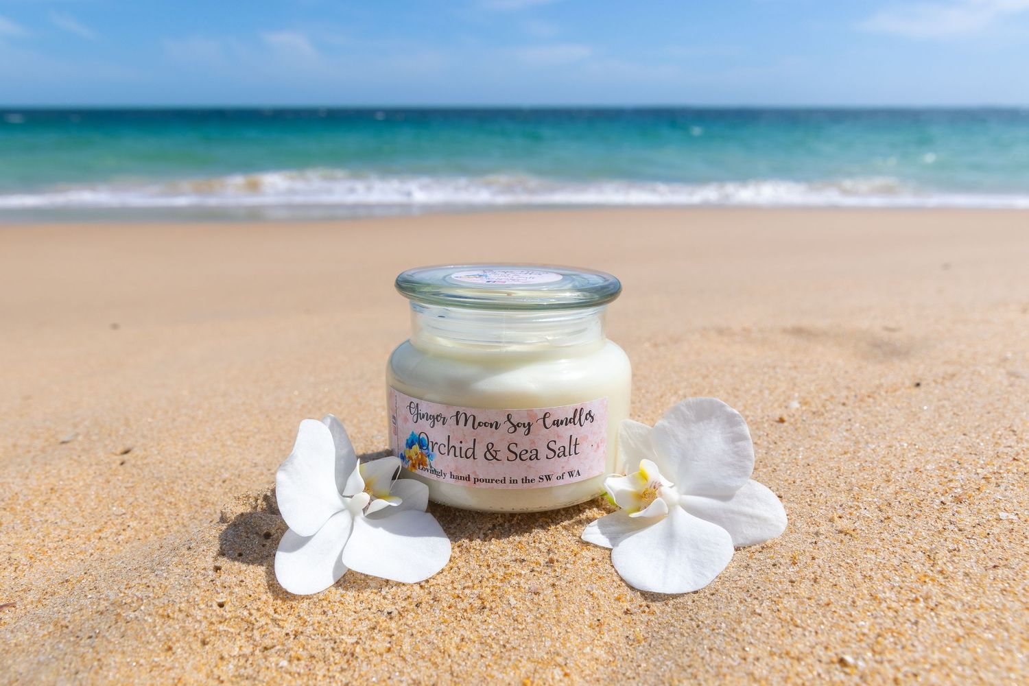 A glass jar of sea salt surrounded by white seashells sits on a sandy beach with turquoise ocean waves in the background.
