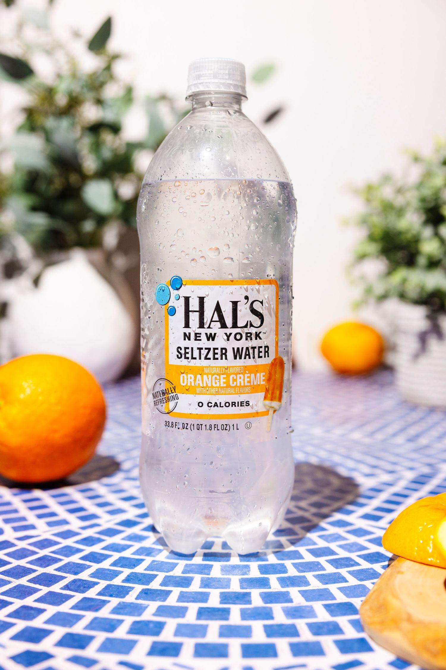 A bottle of Hal's New York Orange Creme Seltzer Water surrounded by fresh oranges on a blue tile surface.