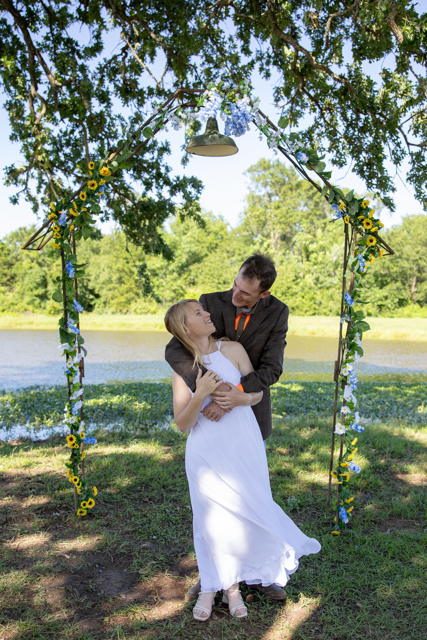 A romantic outdoor wedding scene under a floral arch by a lake with a couple embracing in formal attire.