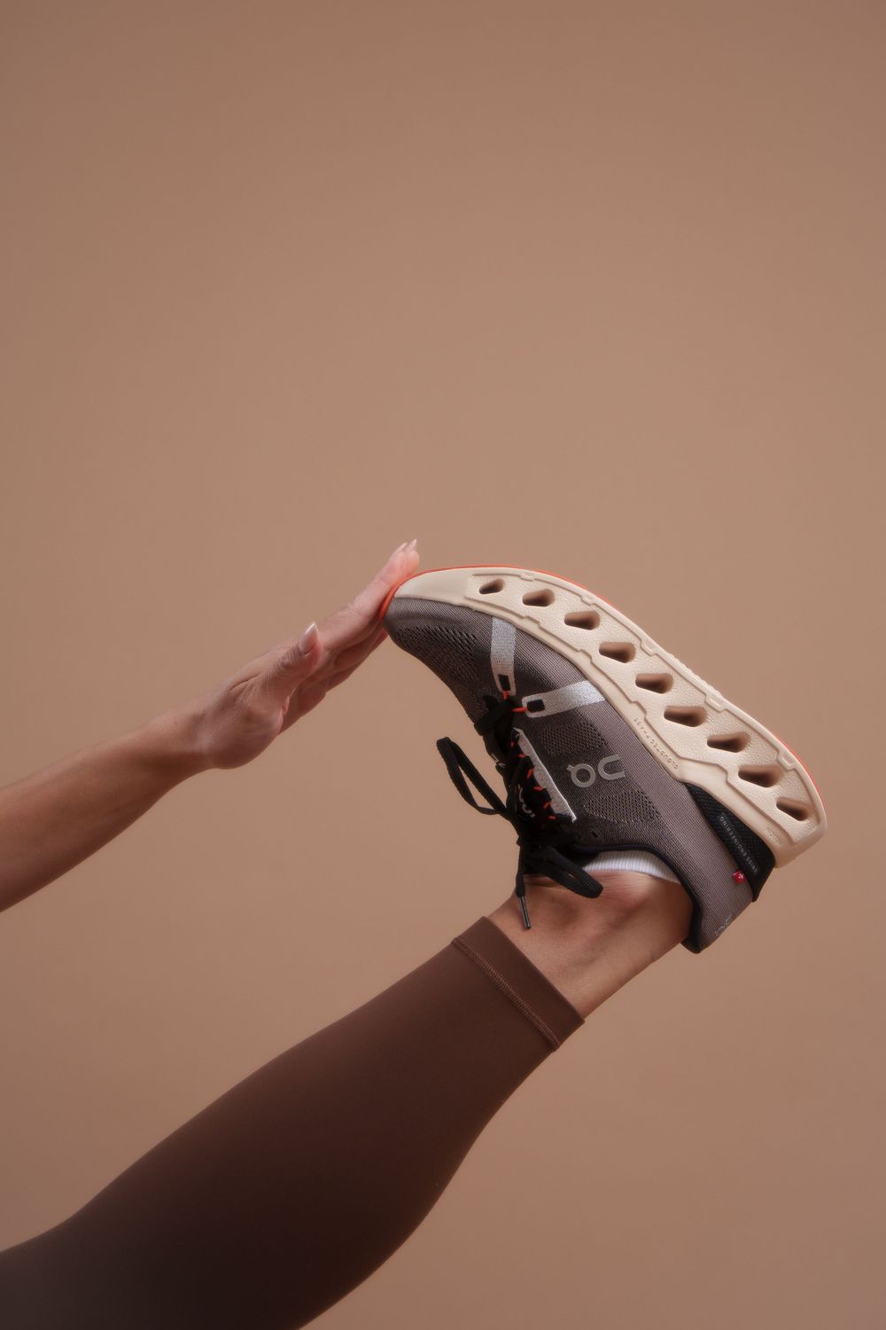Hand reaching out to touch a gray sneaker with unique perforated sole design against a neutral brown background.