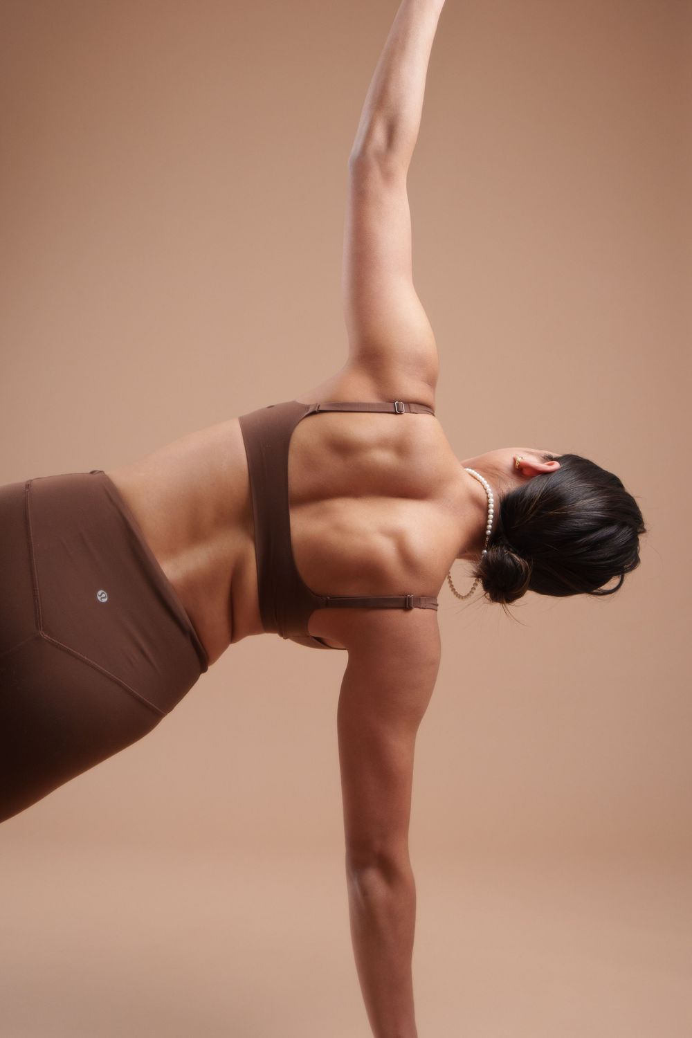 Athletic figure performing a side plank yoga pose wearing brown activewear against a neutral backdrop.
