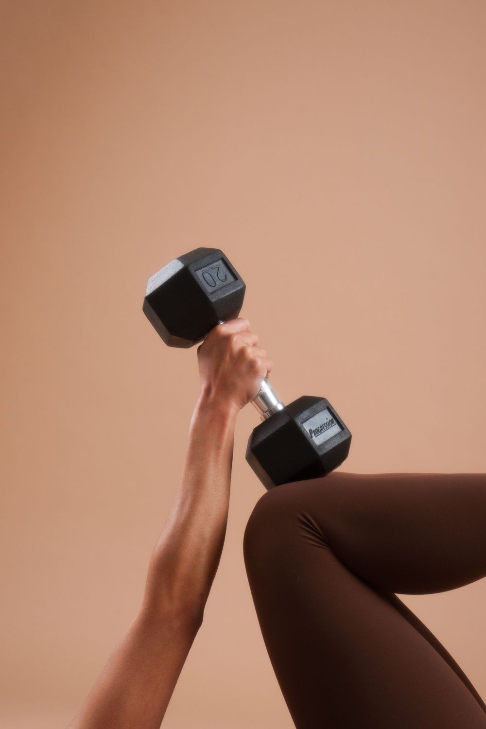 Hand holding a black dumbbell against a peachy brown background while wearing brown leggings.