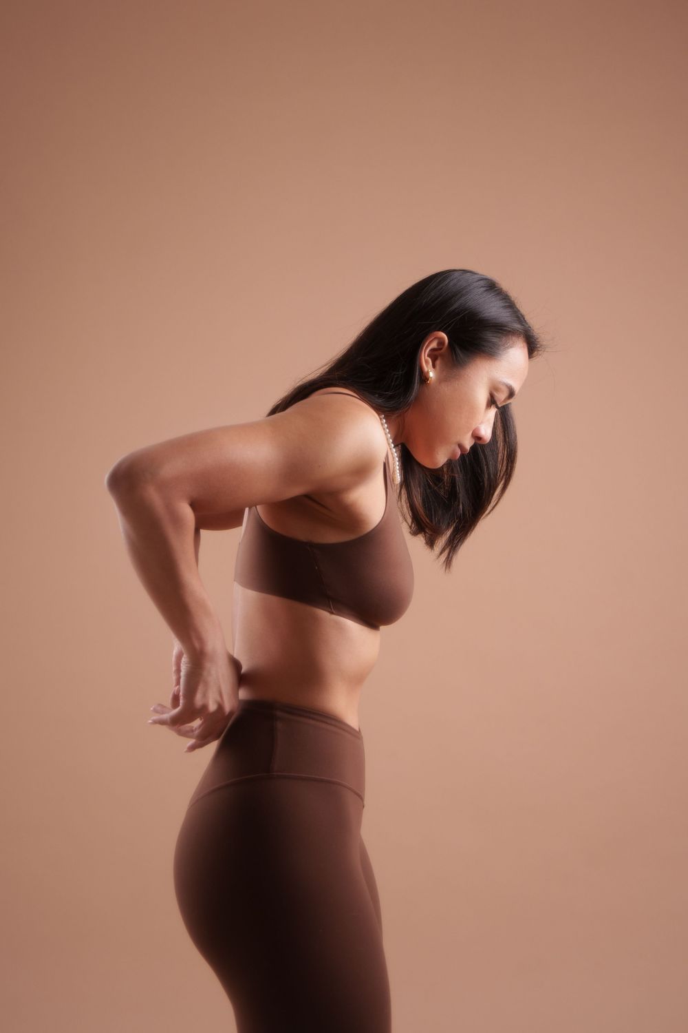 Athletic woman in brown workout attire stretches during fitness routine against beige background.