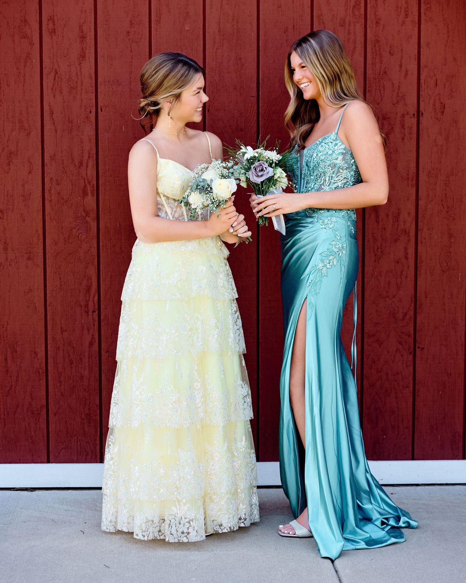 Two friends in elegant prom gowns (yellow lace and turquoise satin) facing each other, smiling and holding bouquets. Outdoor prom portraits.