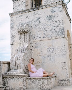 A woman in a pink dress reclines on a white stone ledge against a weathered architectural backdrop.
