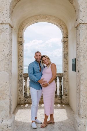A couple poses under white stone arches with ocean views in the background.