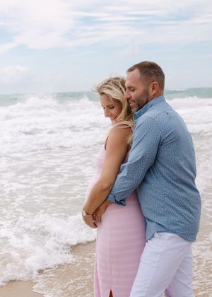A couple embraces on a sandy beach with crashing waves behind them.
