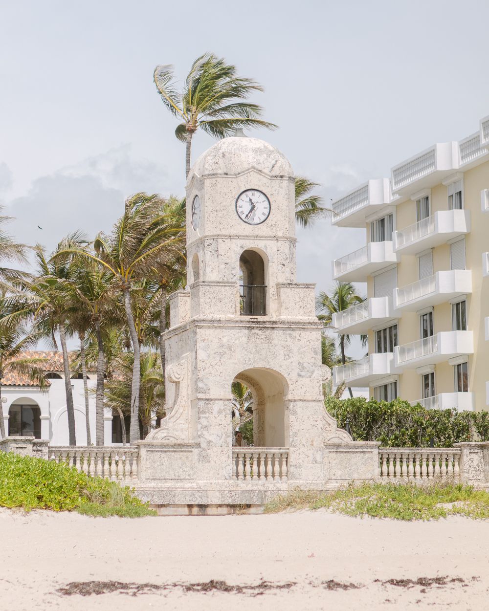 Historic stone clock tower and white building along a palm-lined beach in Palm Beach, Florida on a windy day.