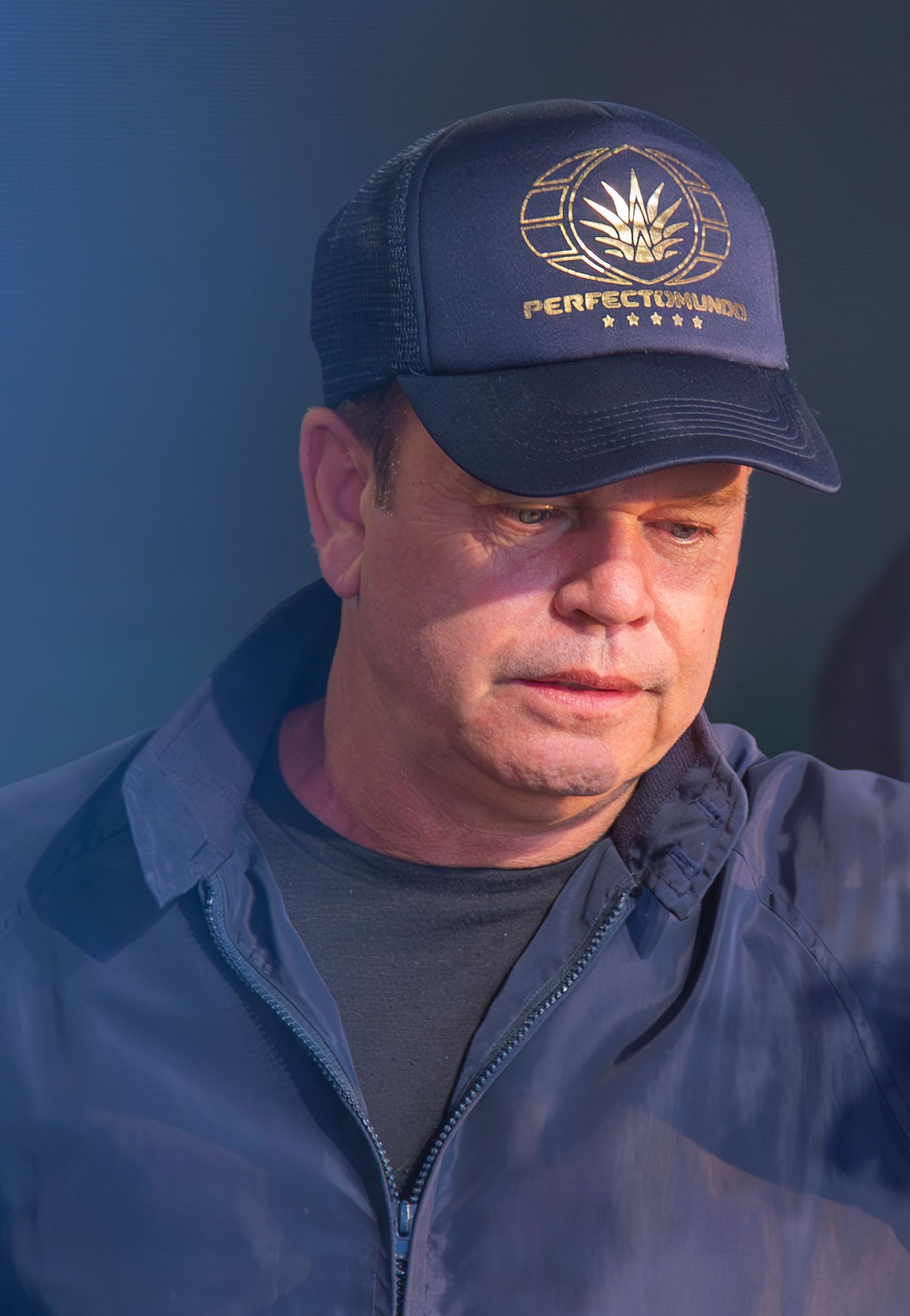 Paul Oakenfold wearing a navy blue trucker hat with a gold embroidered logo and a dark jacket outdoors.