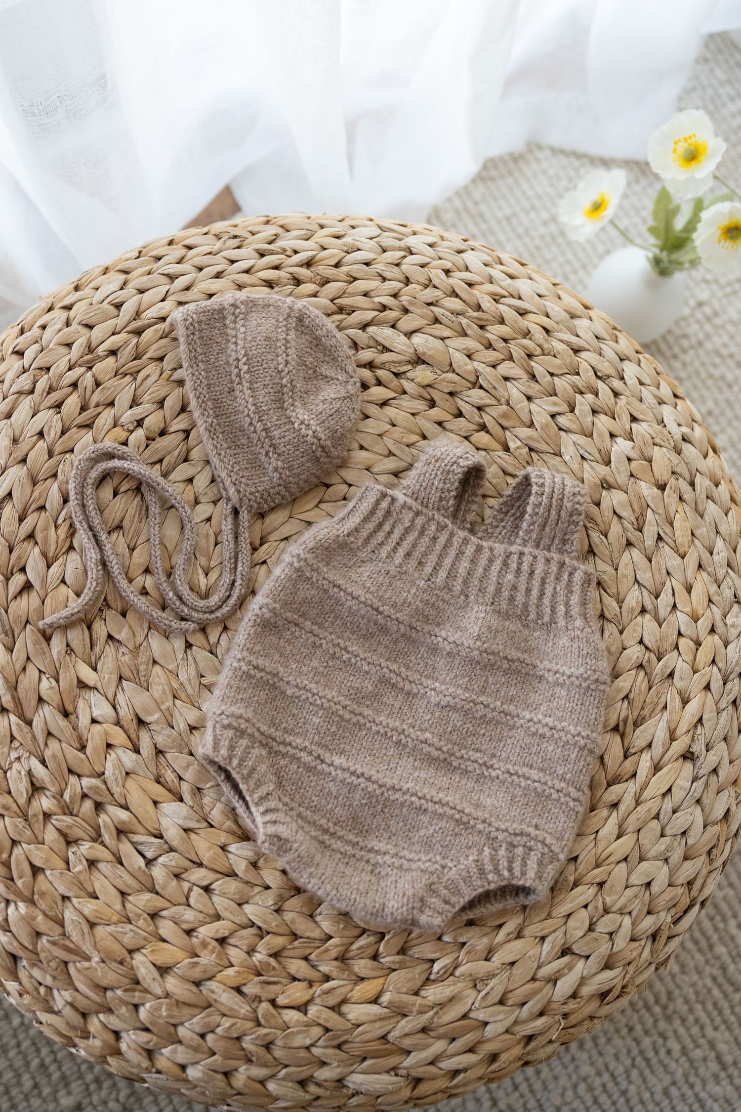 Handmade knitted baby romper and bonnet in neutral beige color displayed on woven straw placemat with daisies.