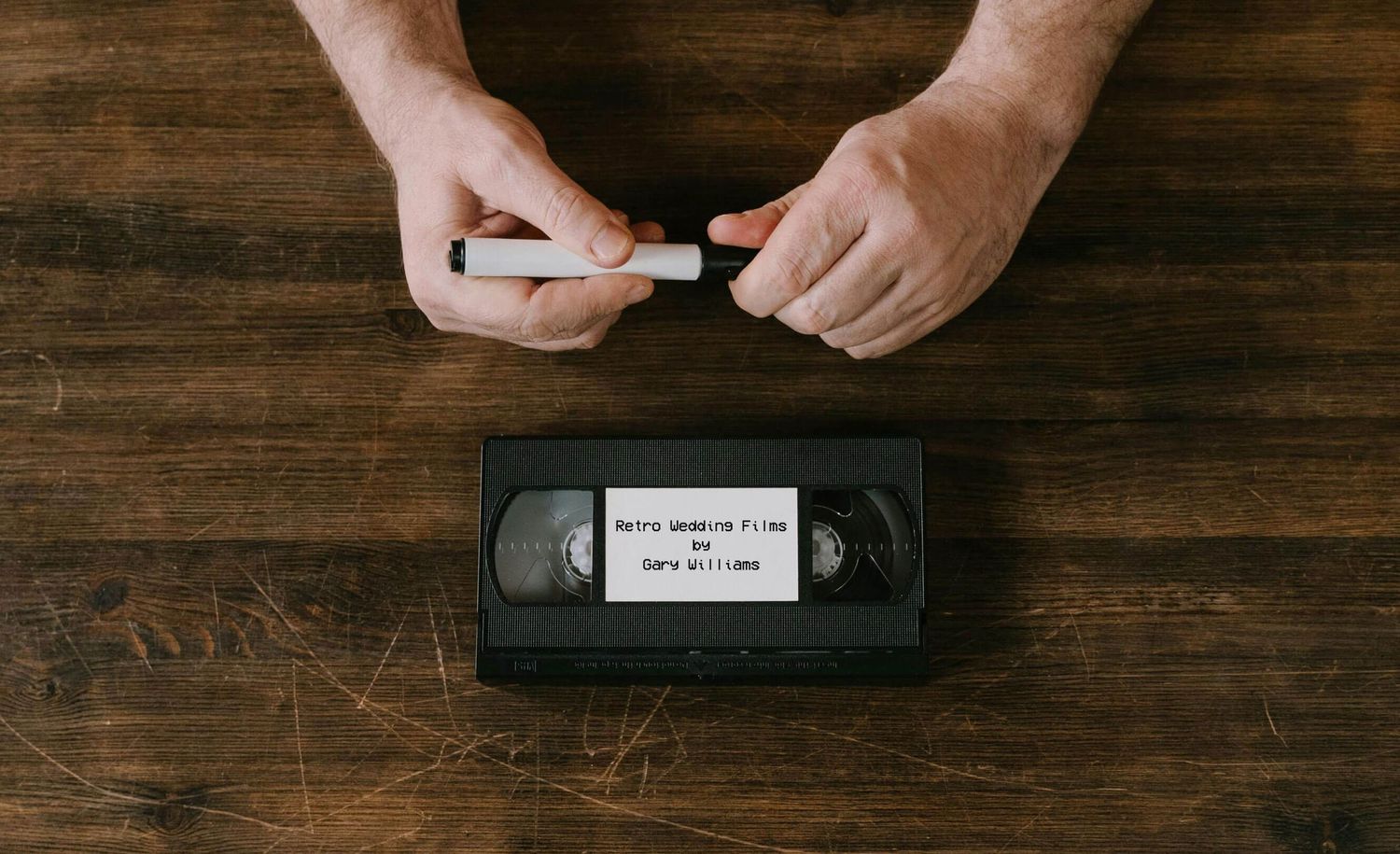 Hands peeling a white sticker label while a VHS tape sits on a dark wooden surface.