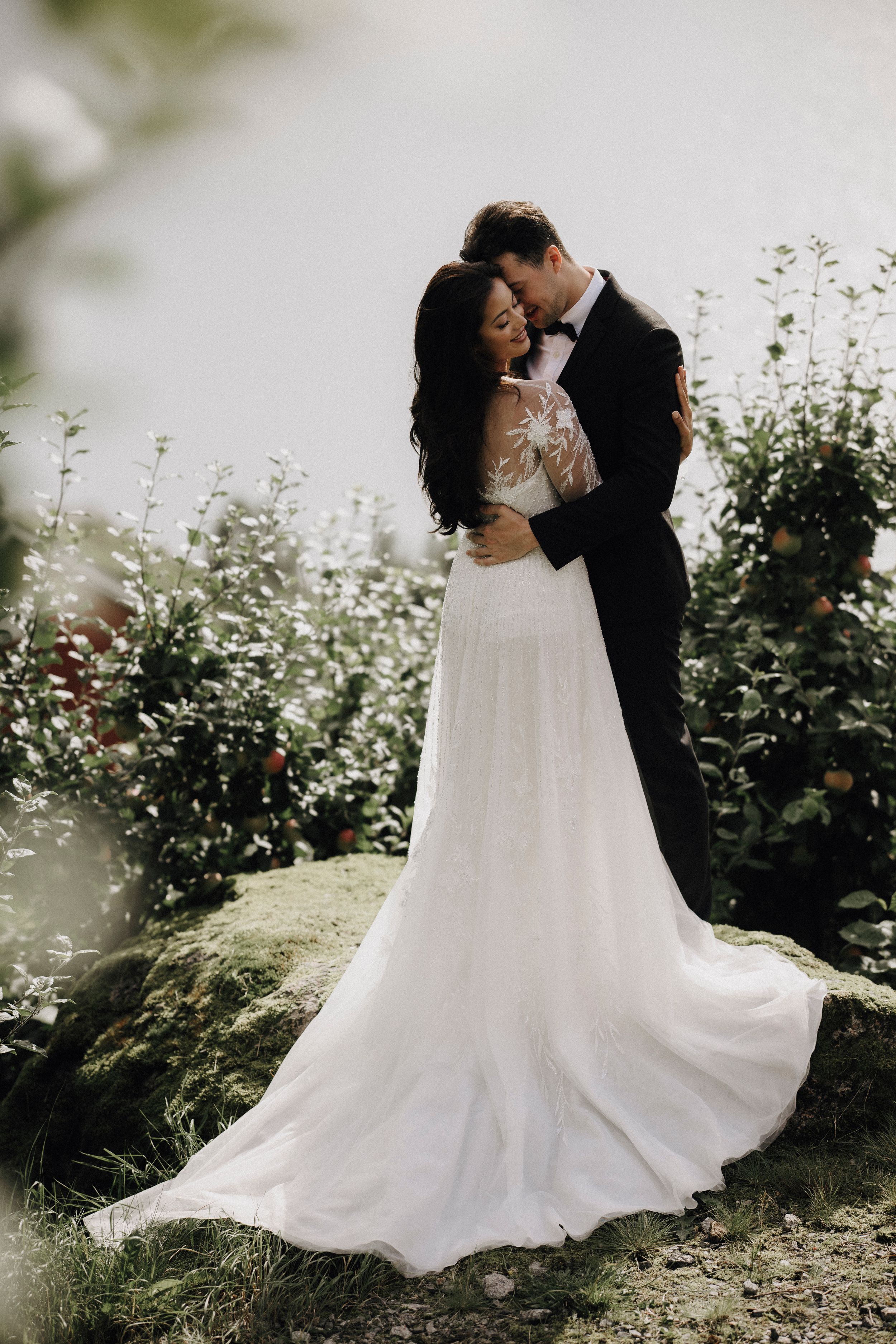 Romantic wedding couple embraces in a garden setting with flowing white dress and lush greenery in soft sunlight.