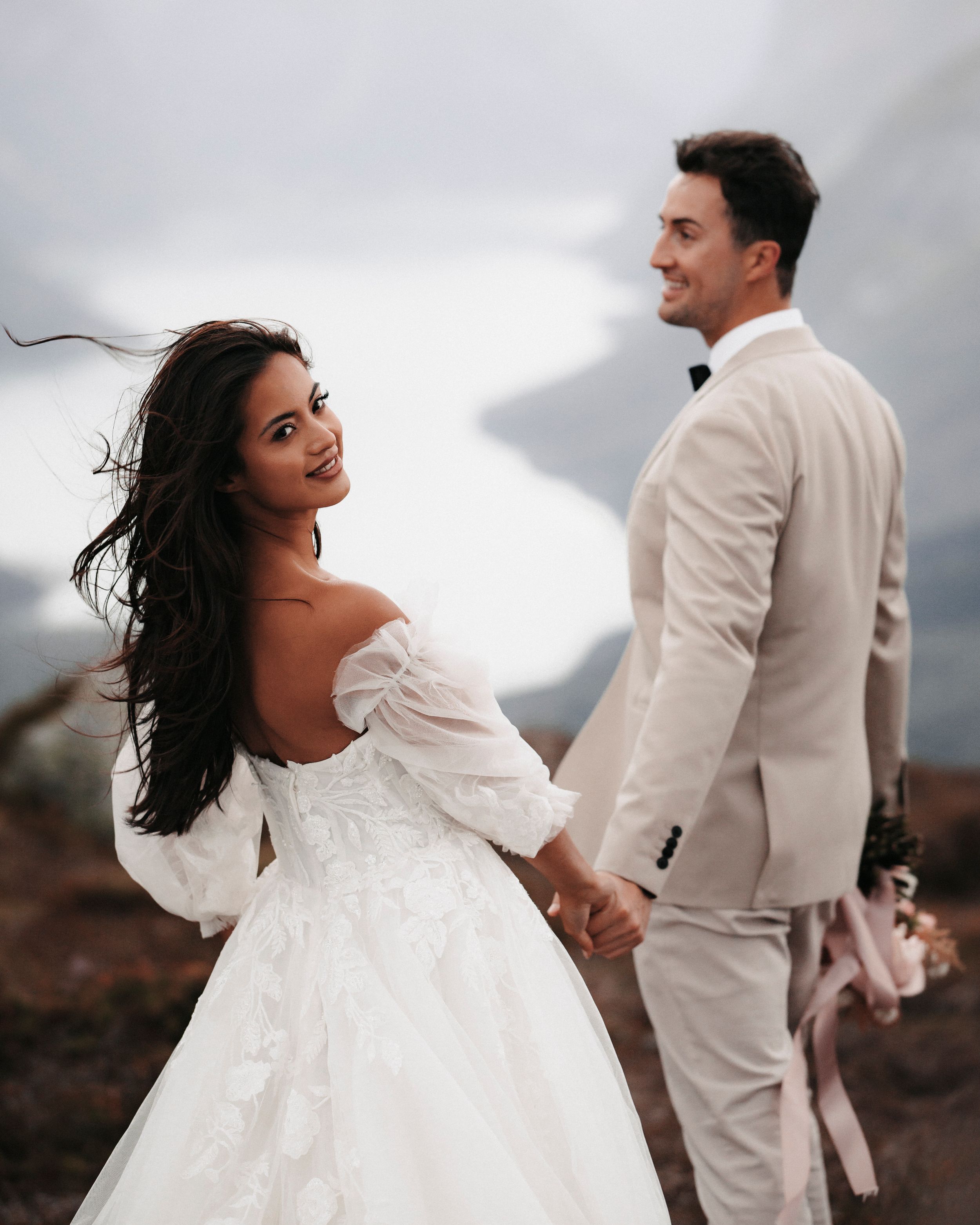 A newlywed couple in white attire pose romantically against a cloudy mountain landscape during their wedding photoshoot.