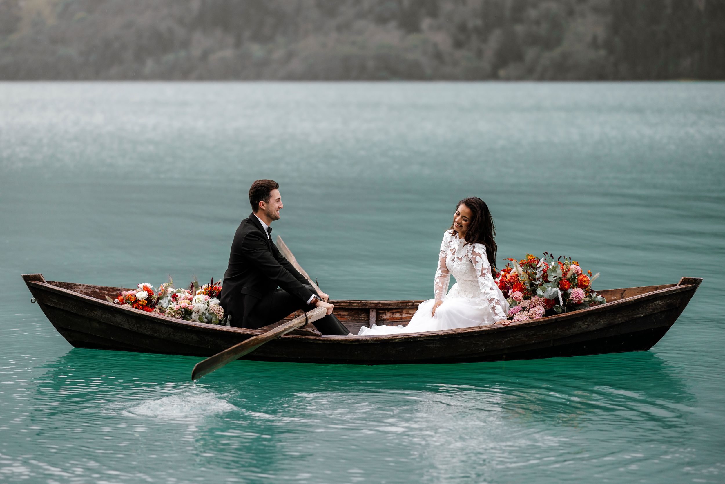 Couple sits in wooden rowboat adorned with floral arrangements on misty turquoise lake during romantic outing.