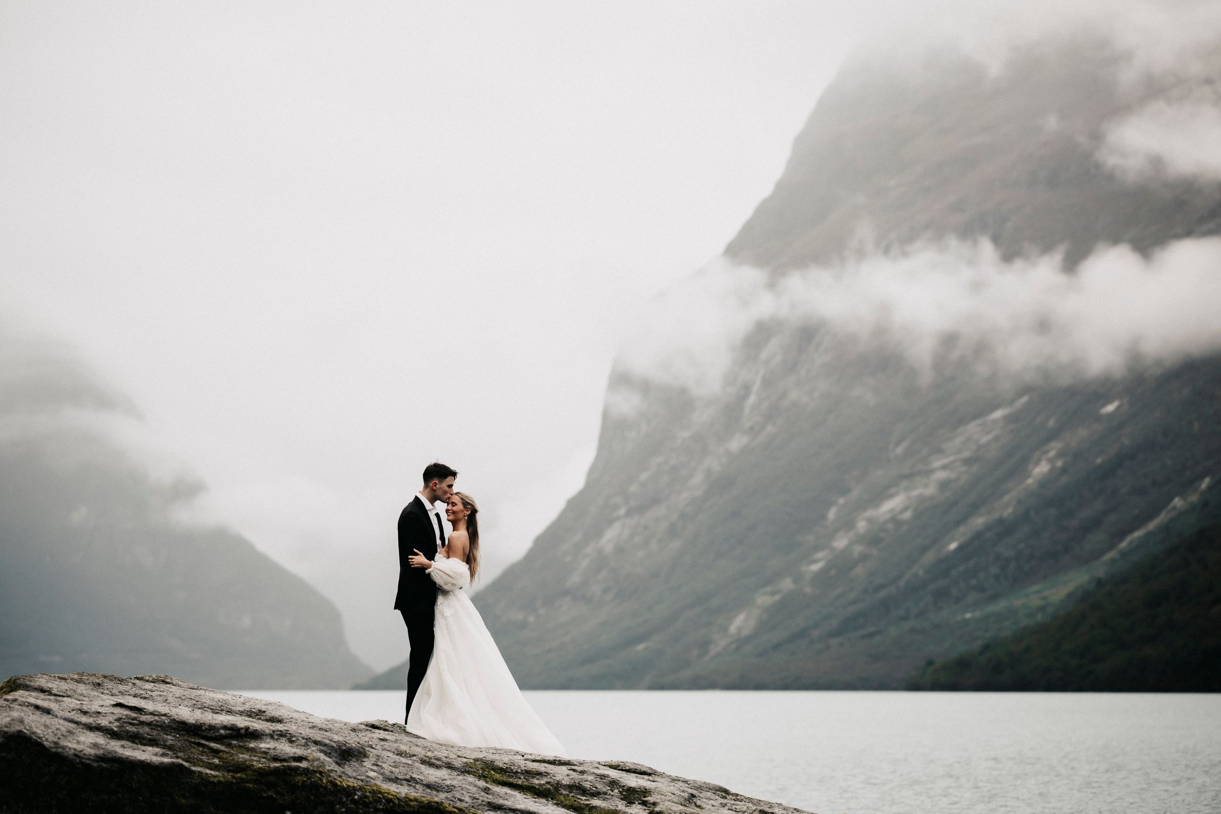A couple in wedding attire embrace on a rocky outcrop overlooking a misty mountainous fjord landscape.