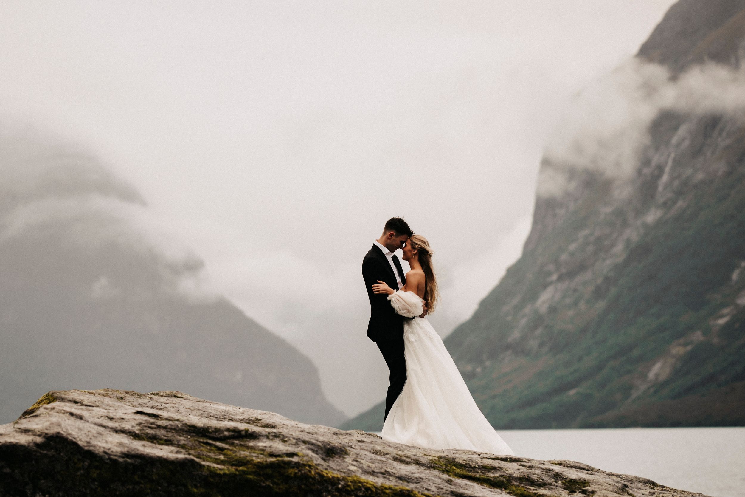 Two people share an intimate moment on a rocky cliff overlooking misty mountains and a fjord in Norway.