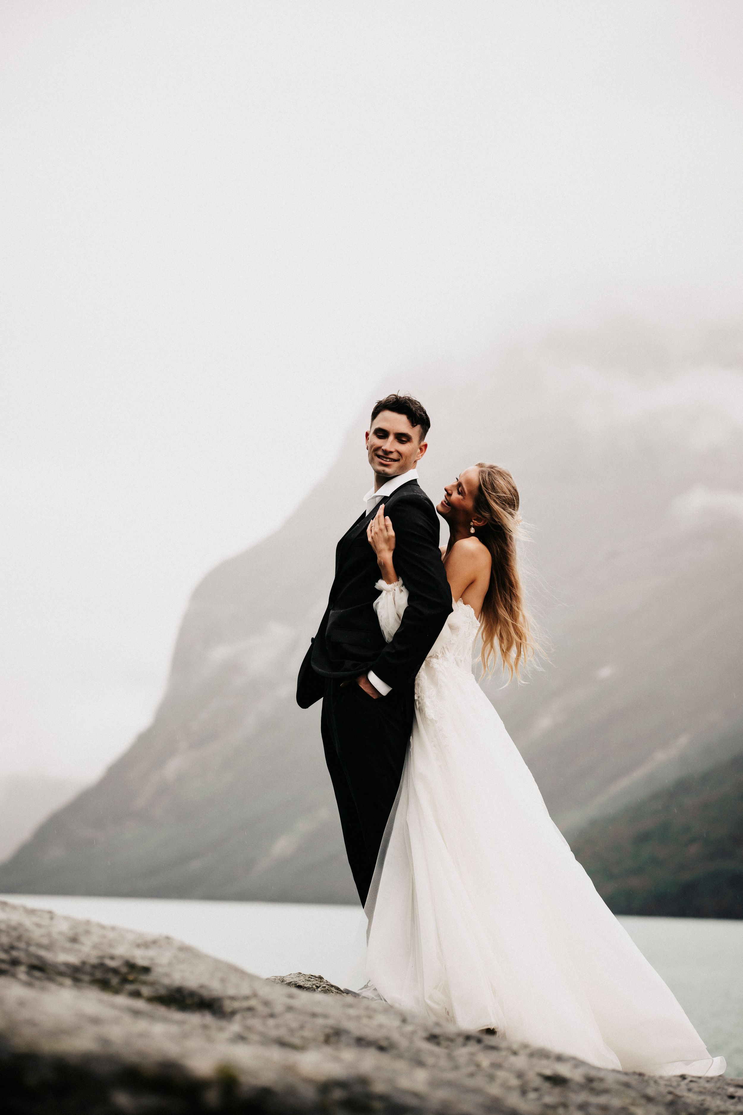 A couple in wedding attire embrace on a cliff edge overlooking a misty mountain lake on their special day.