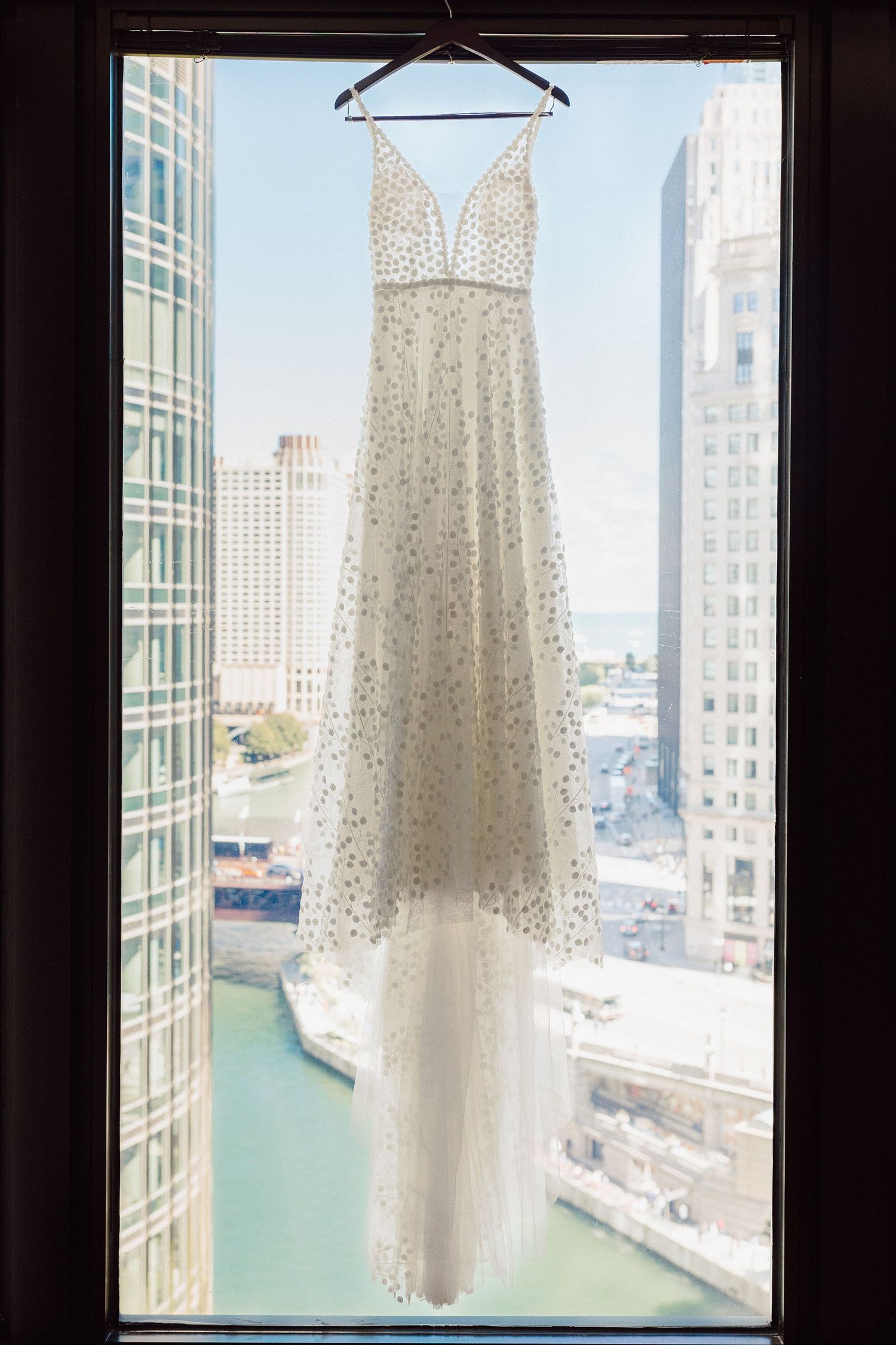 A white beaded wedding dress hanging in front of a window overlooking a city skyline.