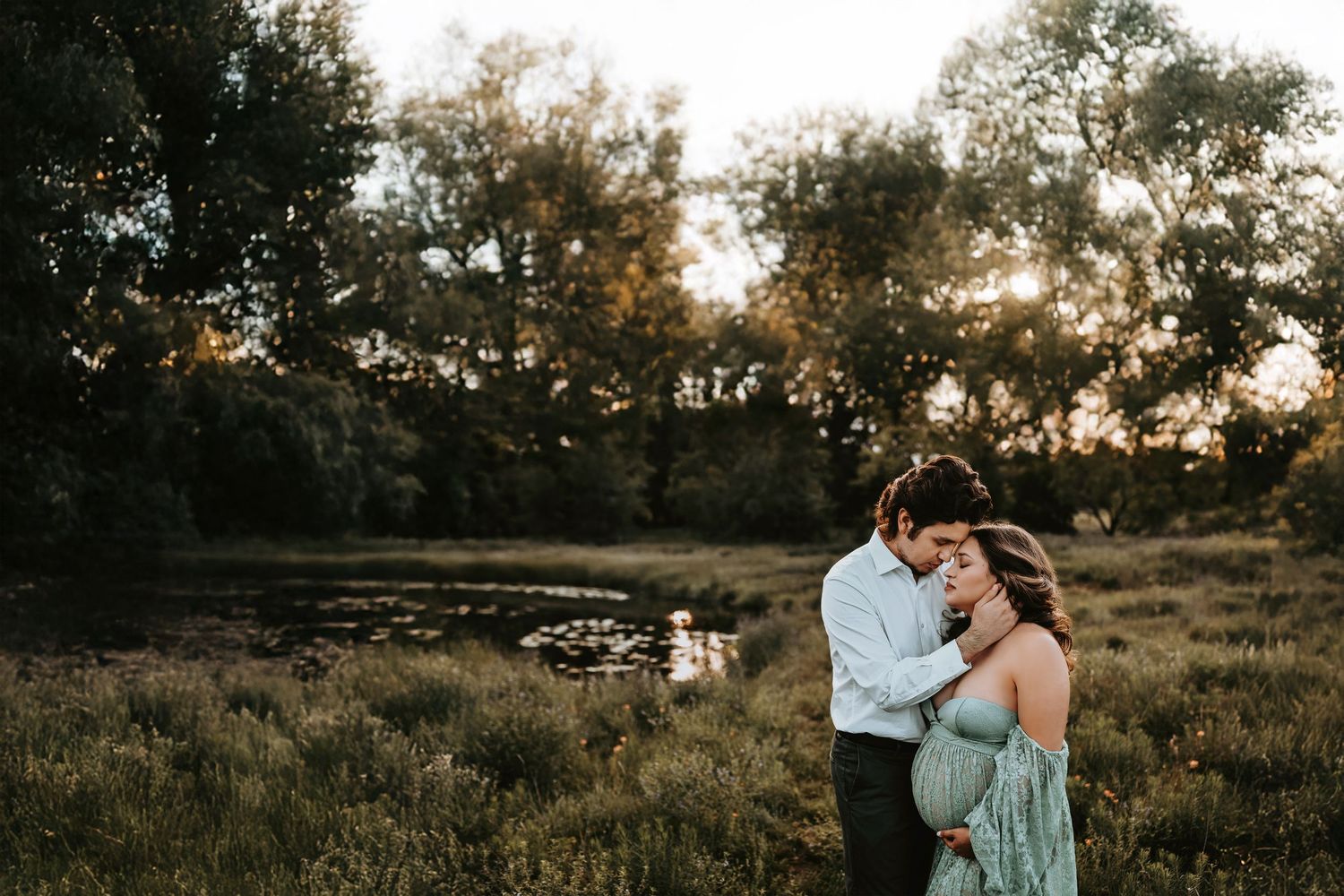 mother and father to be pose lake side in a field at sunset as they embrace for their maternity photos with april pinto photography