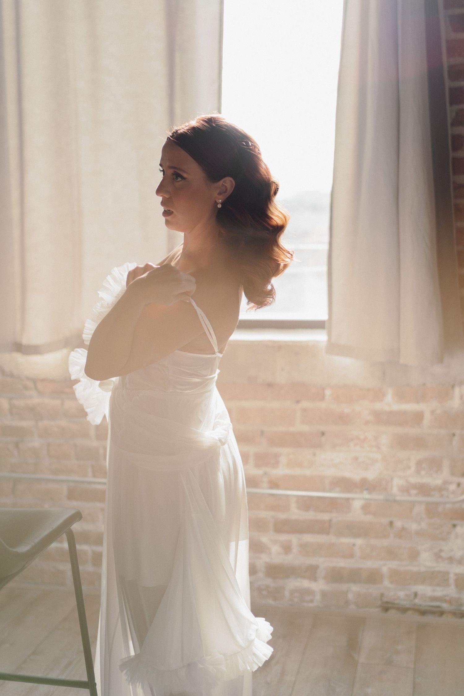 Ethereal bridal portraits by window with exposed brick wall and white curtains.