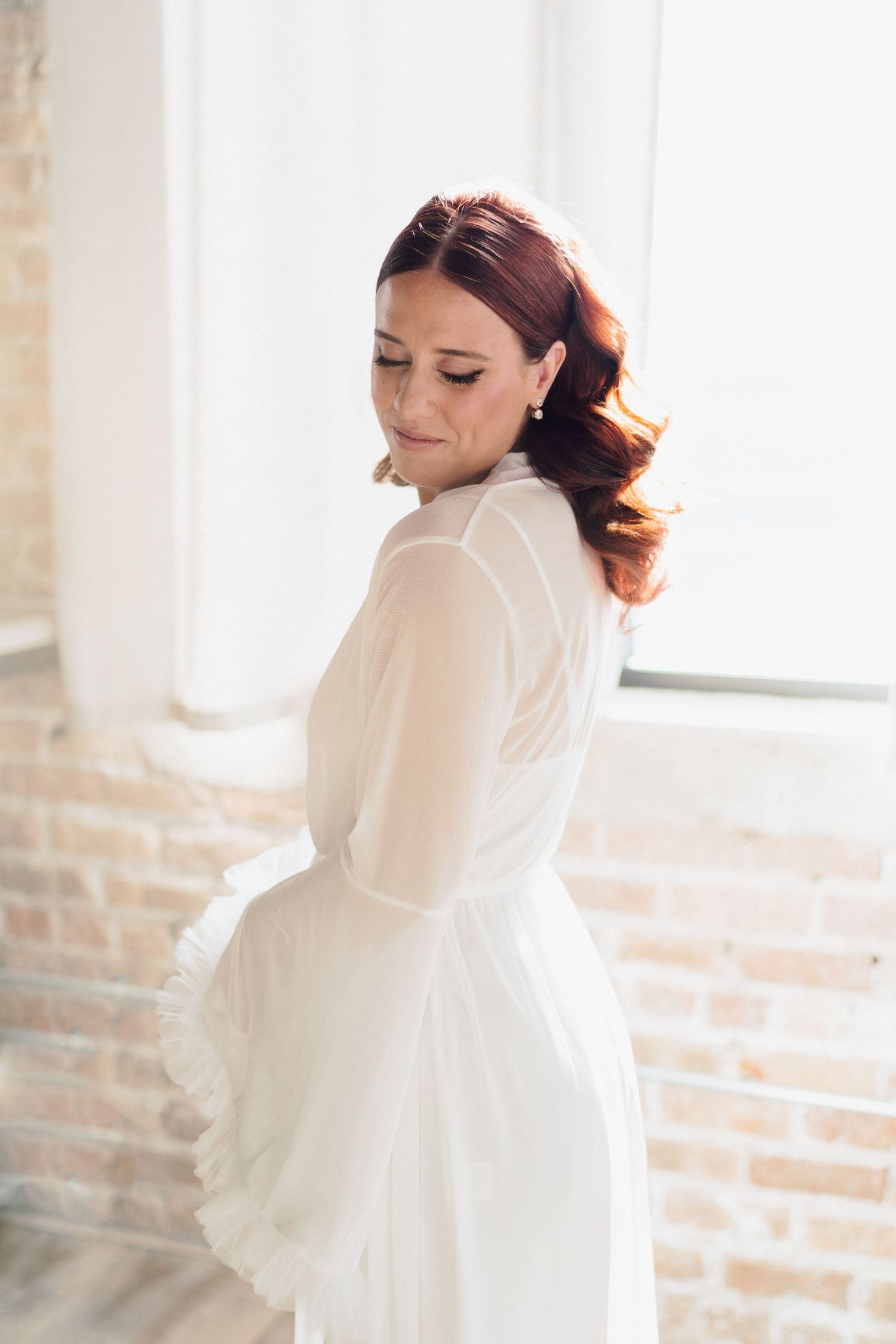 Elegant white wedding dress with sheer details shown in a bright studio setting with exposed brick walls.