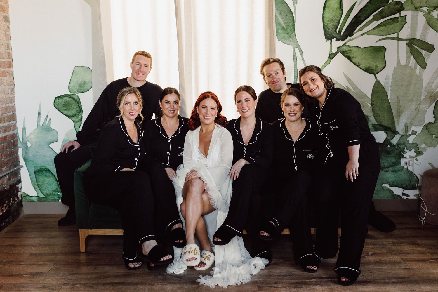 Wedding party poses together in formal attire against a modern studio backdrop with tropical plants.