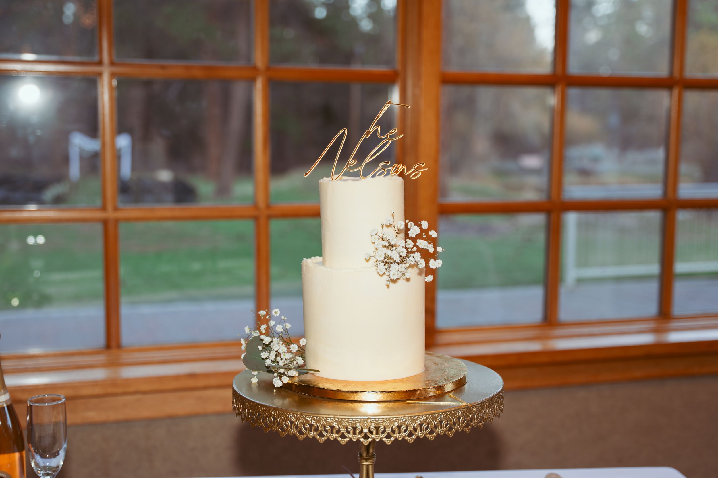 Elegant two-tier white wedding cake with baby's breath flowers on gold cake stand displayed by window.