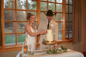Couple cutting their elegant white wedding cake by large windows in rustic venue.