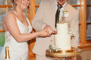 A couple in wedding attire cuts into their elegant white and gold wedding cake on an ornate gold cake stand.