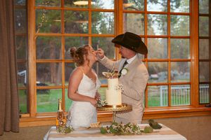 A newlywed couple sharing cake during a wedding reception in a rustic venue with large windows overlooking the outdoors.