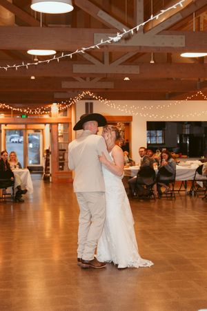 Couple shares first dance under string lights in rustic reception venue with wooden beam ceiling.