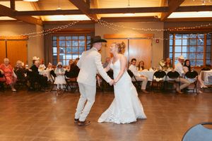 A couple shares their first dance at a wedding reception in a warmly-lit rustic venue with string lights and wooden beams.