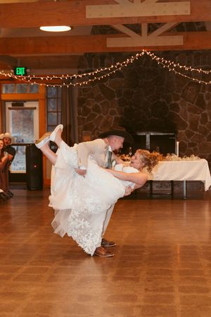 Couple performs dramatic dance move during wedding reception in a lodge with string lights and wooden beams.