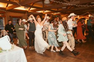 Wedding party dances and celebrates on rustic wooden dance floor decorated with string lights at indoor reception.