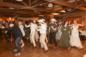 Large group of wedding guests dancing together in rustic reception venue with string lights overhead.