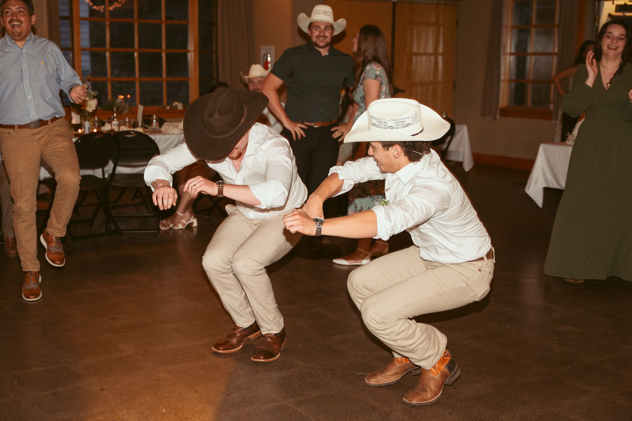 Two people in white outfits and cowboy hats dancing energetically at a party venue with wooden decor.