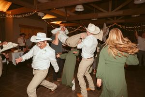 People in cowboy hats and western attire dancing energetically at a rustic indoor venue with string lights.