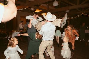 Wedding guests dancing and celebrating at reception with cowboy hats and string lights overhead.