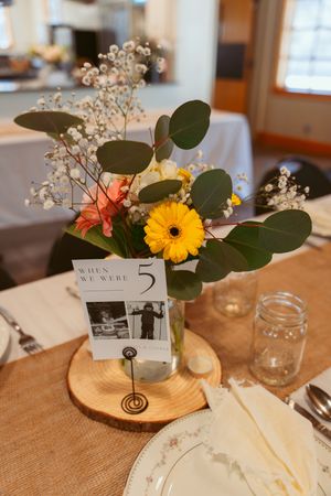 A rustic wedding centerpiece with yellow gerbera daisy, eucalyptus leaves, and table number on a wooden slice.