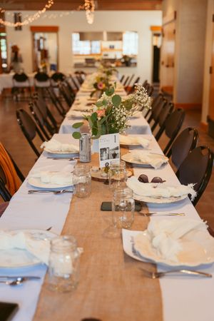 Long rustic wedding reception table with white linens, burlap runner and greenery centerpieces set for dinner.