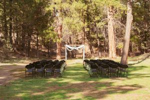 Rustic outdoor wedding ceremony setup with black chairs and white fabric arch in a sunlit forest clearing.