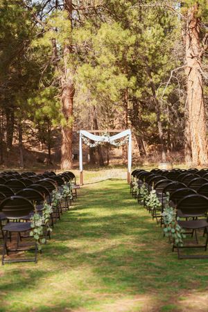 Outdoor wedding ceremony setup with rows of folding chairs and floral arch in a pine forest setting.