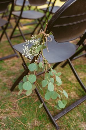A decorative eucalyptus and baby's breath floral arrangement tied to a black folding chair outdoors.